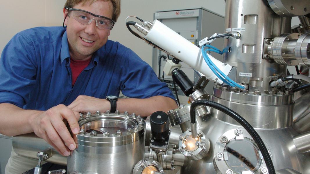 Smiling man in blue shirt and safety goggles amidst equipment.