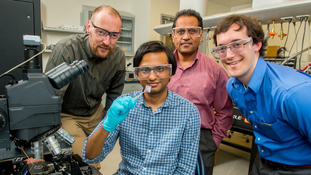 Four goggled men in lab, smiling at camera. One holds small square in tweezers.