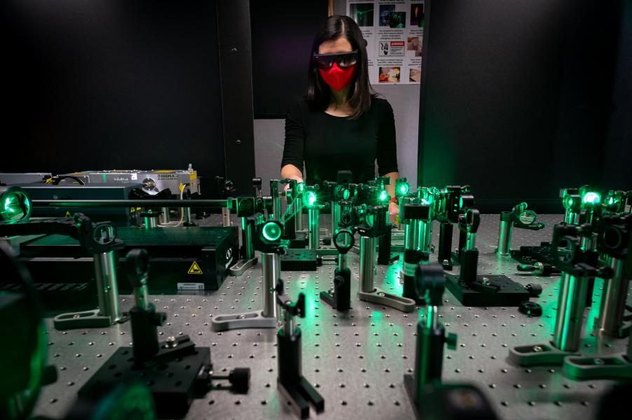 Woman in red face mask and dark glasses looking at perforated table holding many green glowing cylinders.